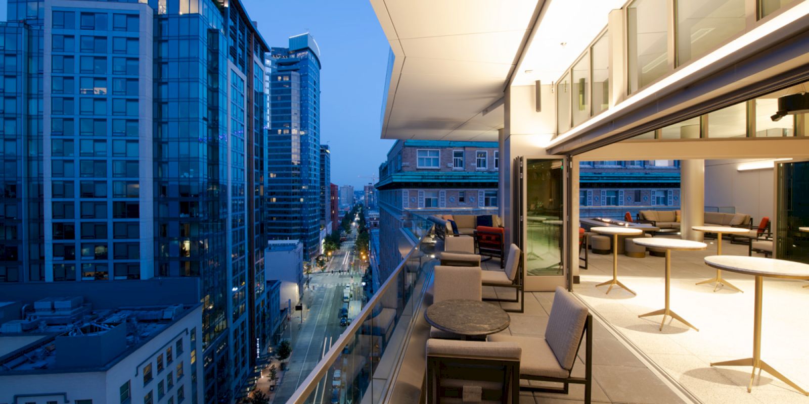 The image shows a modern rooftop terrace with seating, overlooking city buildings as evening falls, creating a cozy urban atmosphere.