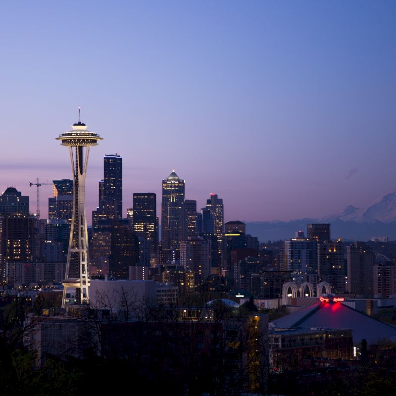 A cityscape at dusk featuring the Space Needle and Seattle skyline with a mountain in the background under a colorful sky.