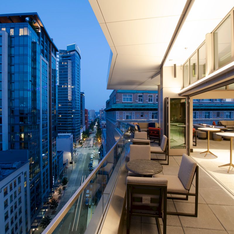 A modern urban balcony at dusk, overlooking a city street with tall glass buildings, featuring outdoor seating and ambient lighting.