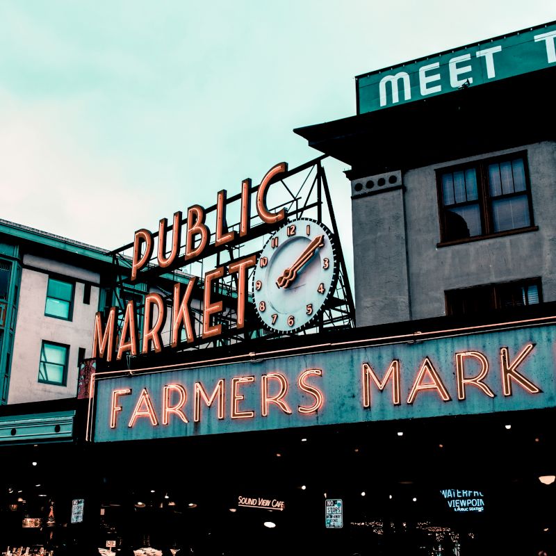 The image shows the Pike Place Market sign, with neon lights displaying "Public Market" and "Farmers Market" against a cityscape background.