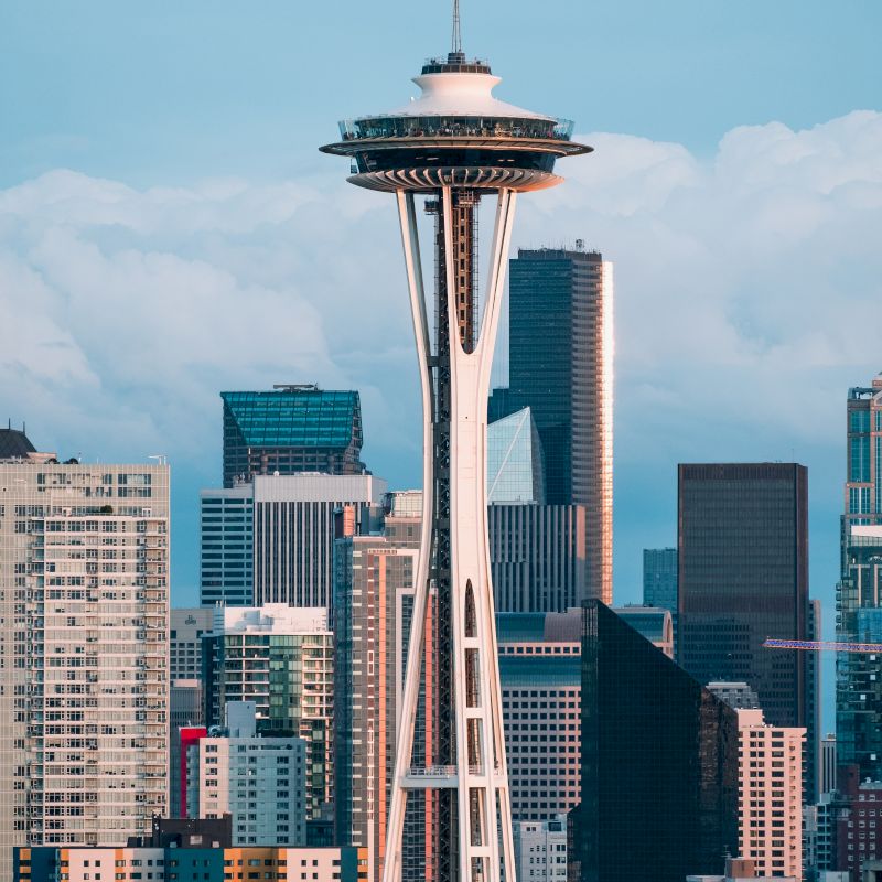 The image shows the Space Needle amidst the Seattle skyline, with various skyscrapers and buildings against a cloudy sky backdrop.
