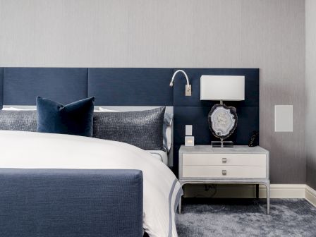 A modern bedroom with a navy headboard, white bedside table, lamp, and decorative items, featuring a neatly made bed with pillows.