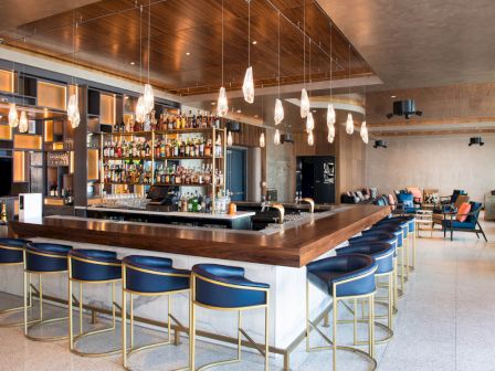 A modern bar with blue stools, a wooden counter, pendant lights, and shelves stocked with bottles. Bright and inviting atmosphere.