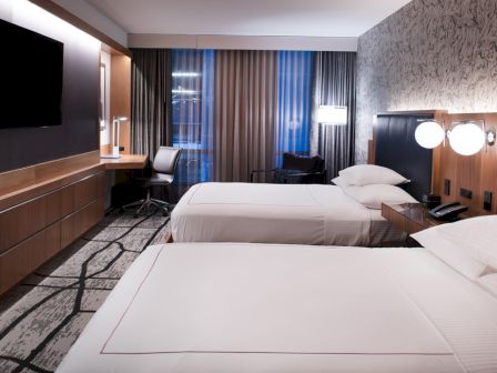 The image shows a modern hotel room with two beds, a wall-mounted TV, a work desk, and stylish lighting. The decor is contemporary and sleek.