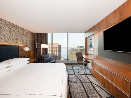 A modern hotel room features a large bed, a desk with a chair, a flat-screen TV, and a window with a city view, creating a cozy space.
