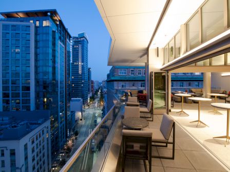 A modern rooftop terrace features seating and tables, overlooking a cityscape with tall buildings during twilight.