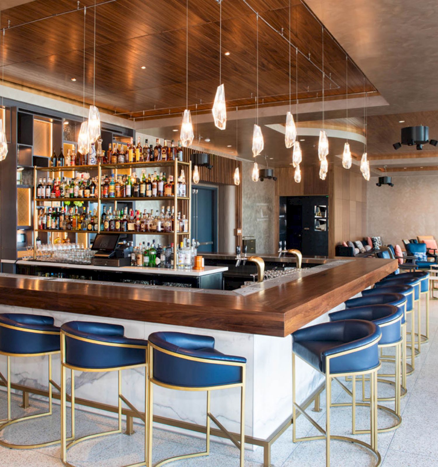 The image shows a modern bar with a central counter, blue bar stools, pendant lights, and a well-stocked shelf, in a stylish setting.