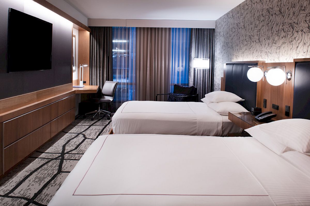 A modern hotel room with two beds, a wall-mounted TV, a desk with a chair, and stylish lighting and decor.