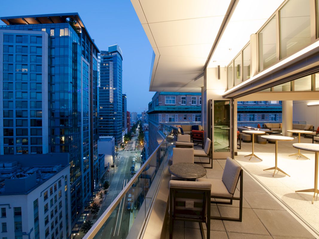A modern rooftop terrace at dusk with seating areas, overlooking a cityscape with tall buildings and street lights visible below.