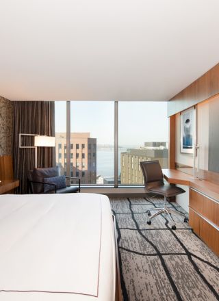 A modern hotel room with a double bed, large TV, desk, and floor-to-ceiling window featuring a city view.