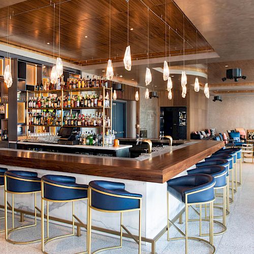 The image shows a modern bar with blue stools, a central counter, shelves with bottles, and pendant lights in a stylish interior.