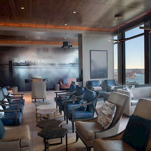 The image shows a stylish lounge with modern seating, art on the wall, and large windows showcasing a scenic view of the water and sky.