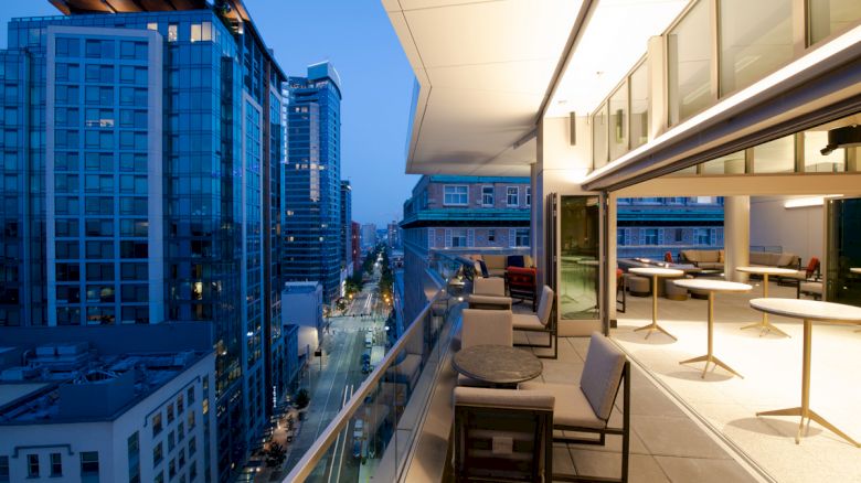A modern outdoor terrace overlooks a cityscape at dusk, with tall glass buildings and a well-lit, stylish seating area.