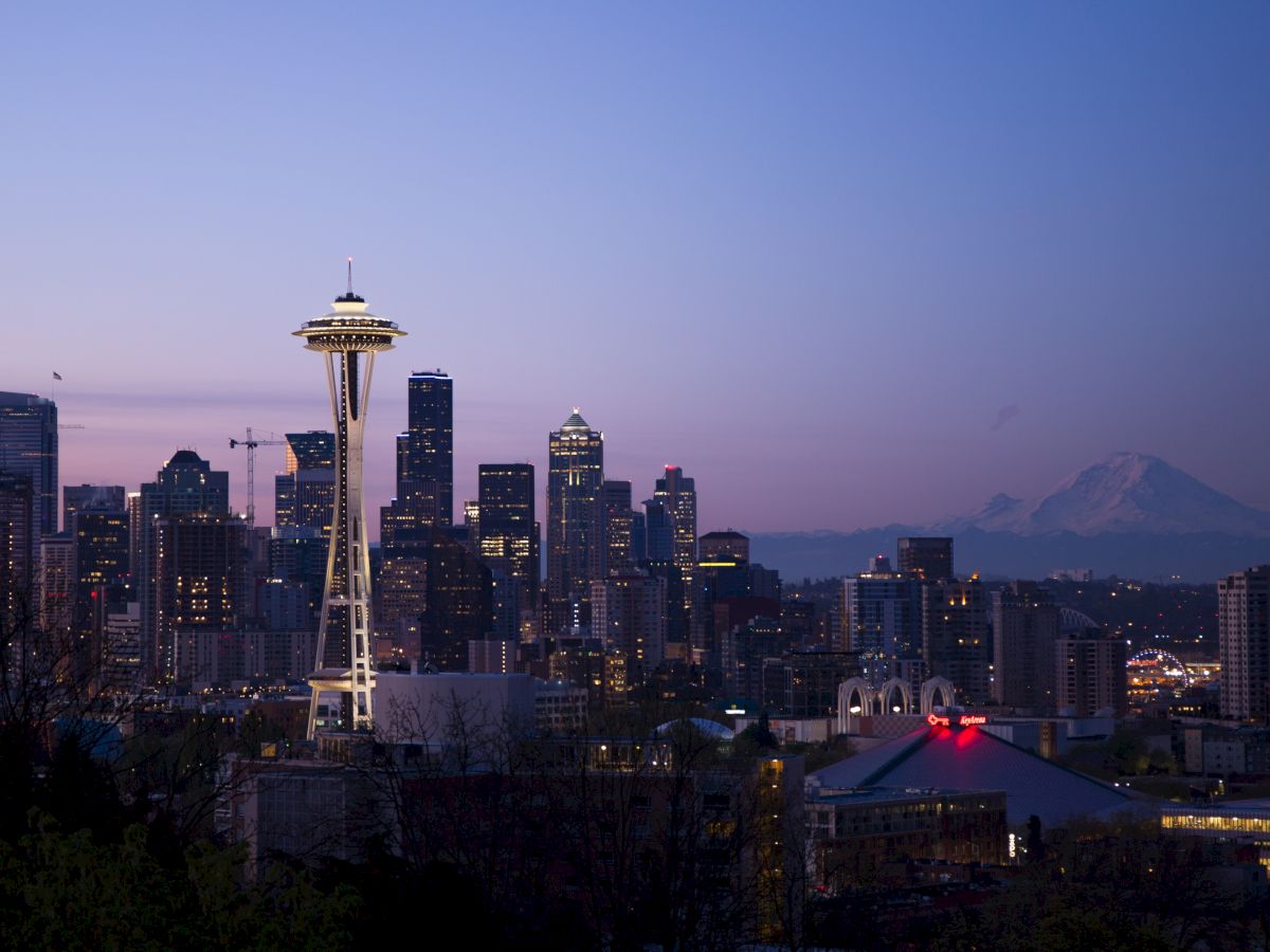 A city skyline at dusk features the Space Needle, with a mountainous backdrop under a pink and purple sky.