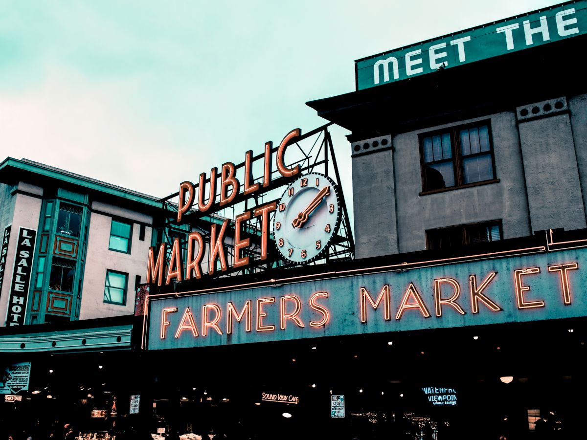 The image shows the iconic entrance sign of a public market with a clock and neon "Farmers Market" lettering, set against an urban backdrop.