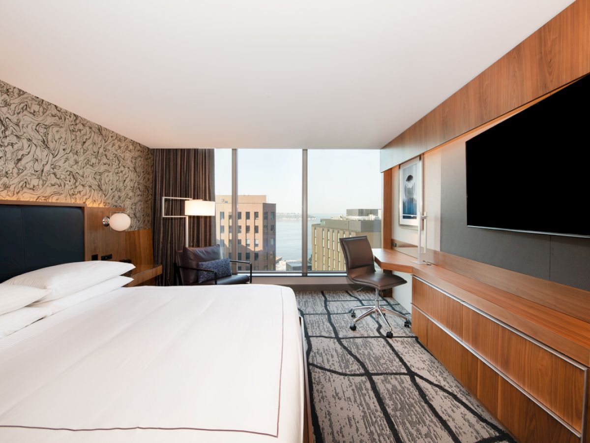A modern hotel room with a large bed, wall-mounted TV, desk, and a window with a city view.
