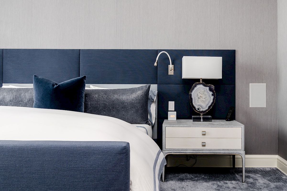 A modern bedroom with a blue headboard, crisp bedding, nightstand, lamp, framed photo, and wall sconce, all in a minimalist style.