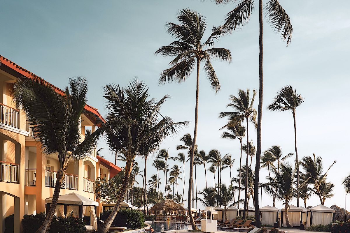 A tropical resort scene with palm trees, a swimming pool, and lounge chairs beside a building with balconies under a clear sky.