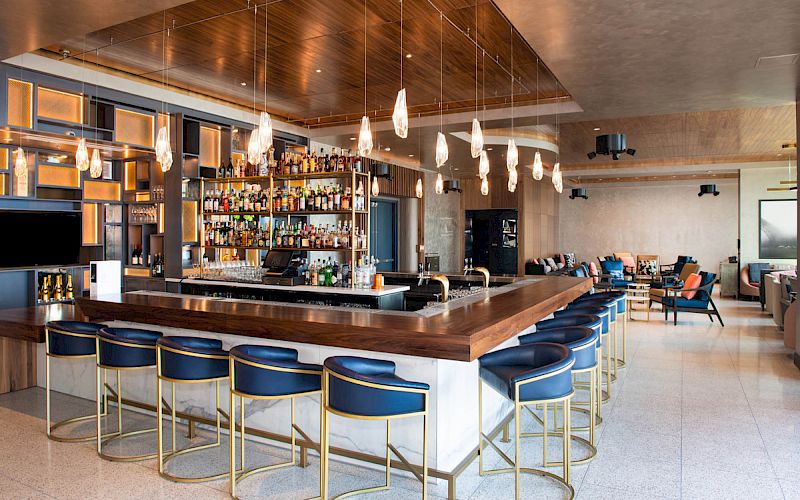 The image shows a modern bar with a wooden counter, blue stools, hanging lights, and shelves stocked with bottles.