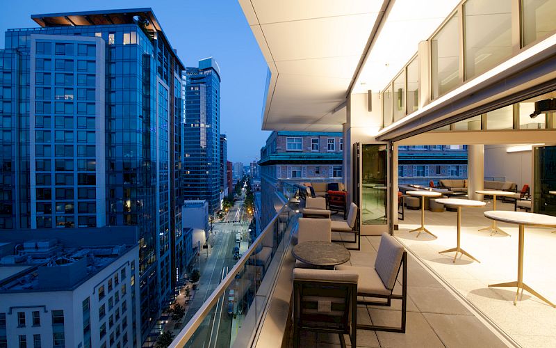 A modern balcony overlooks a cityscape at dusk, with seating and tables arranged for leisure and dining.
