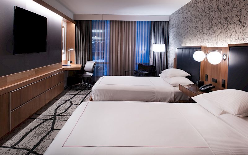 A modern hotel room with two beds, a large TV, a work desk, and ambient lighting. Carpeted floor and patterned curtains complete the design.