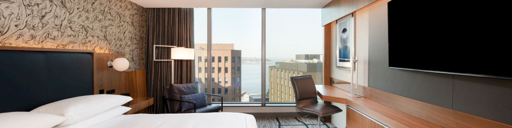 A modern hotel room with a large bed, TV, desk, and chair, featuring a city view through a window.