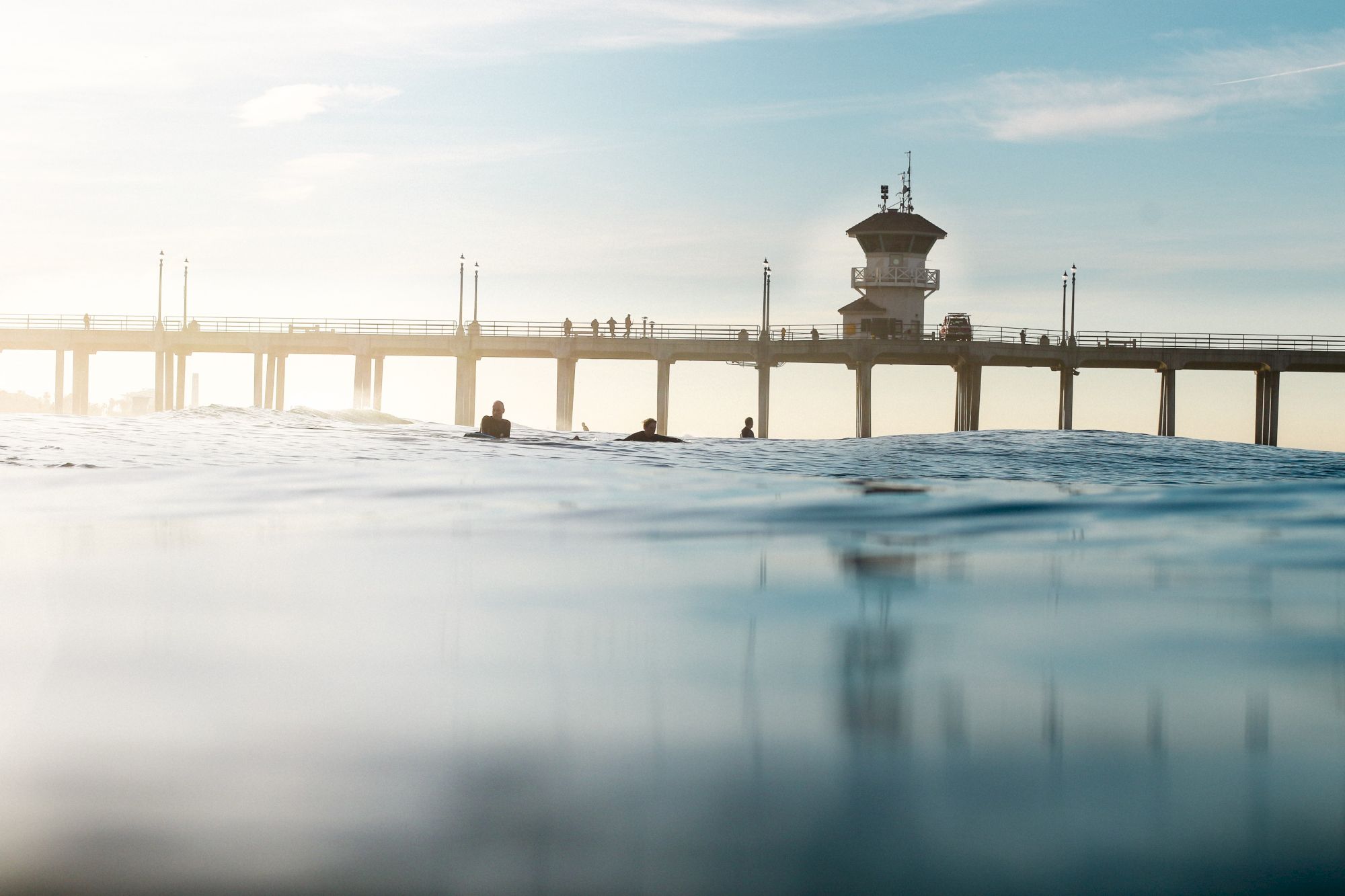 A pier extends into calm water with people walking on it, under a clear sky, viewed from a low angle near the water's surface.